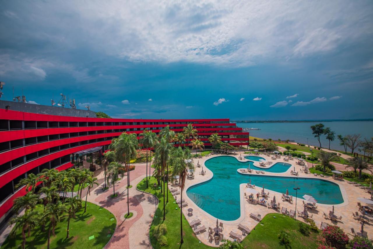 Отель Royal Tulip Brasilia Alvorada Экстерьер фото The photo shows a modern hotel building with a striking red exterior, surrounded by lush greenery. In the foreground, there is a large, creatively shaped swimming pool, with lounge chairs arranged around it. The backdrop features a body of water, pos