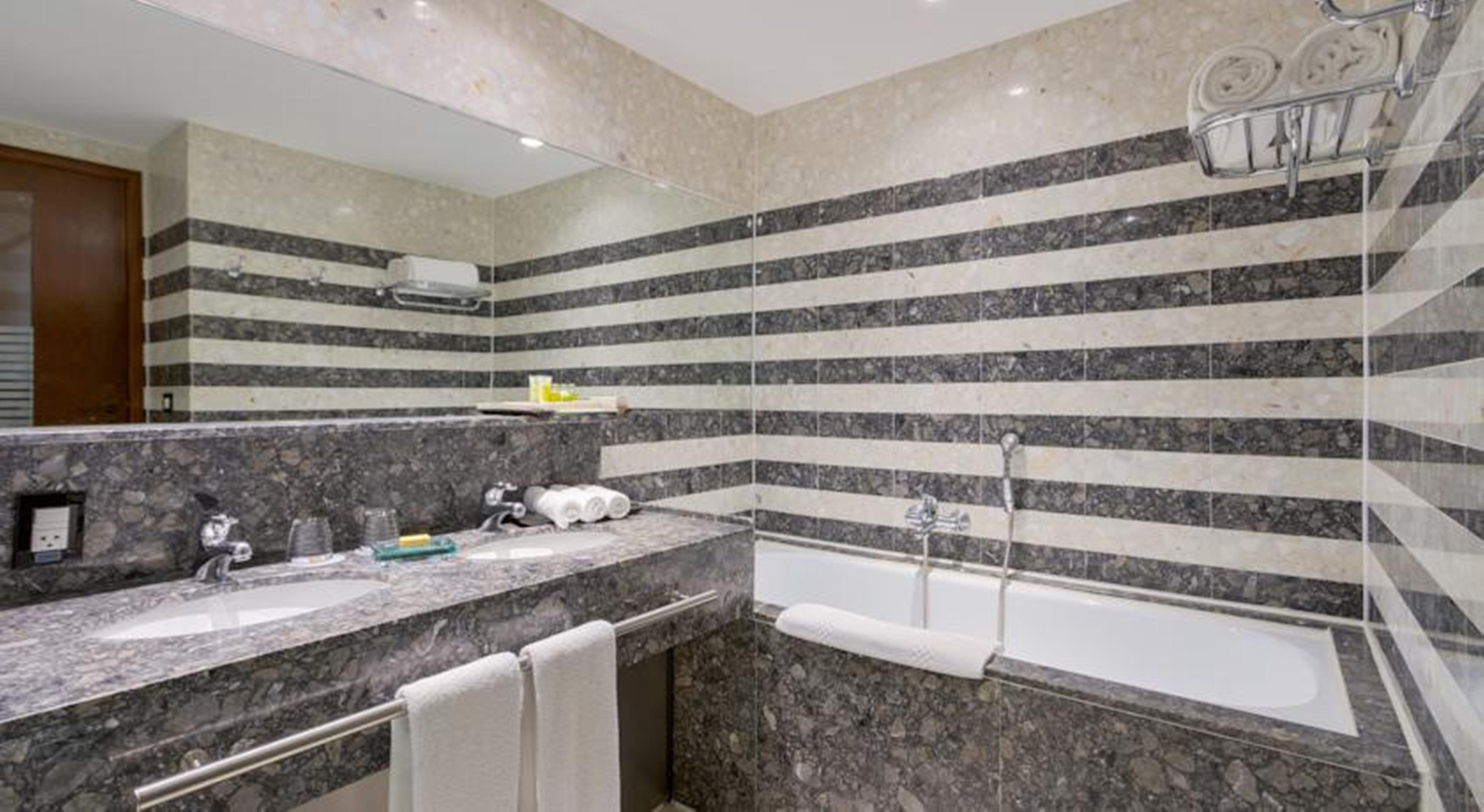 Отель Royal Tulip Brasilia Alvorada Экстерьер фото The photo shows a modern bathroom featuring a combination of marble and stone finishes. The walls are decorated with horizontal stripes in various shades of gray and cream. There is a bathtub with a white exterior, and a showerhead is mounted on the 
