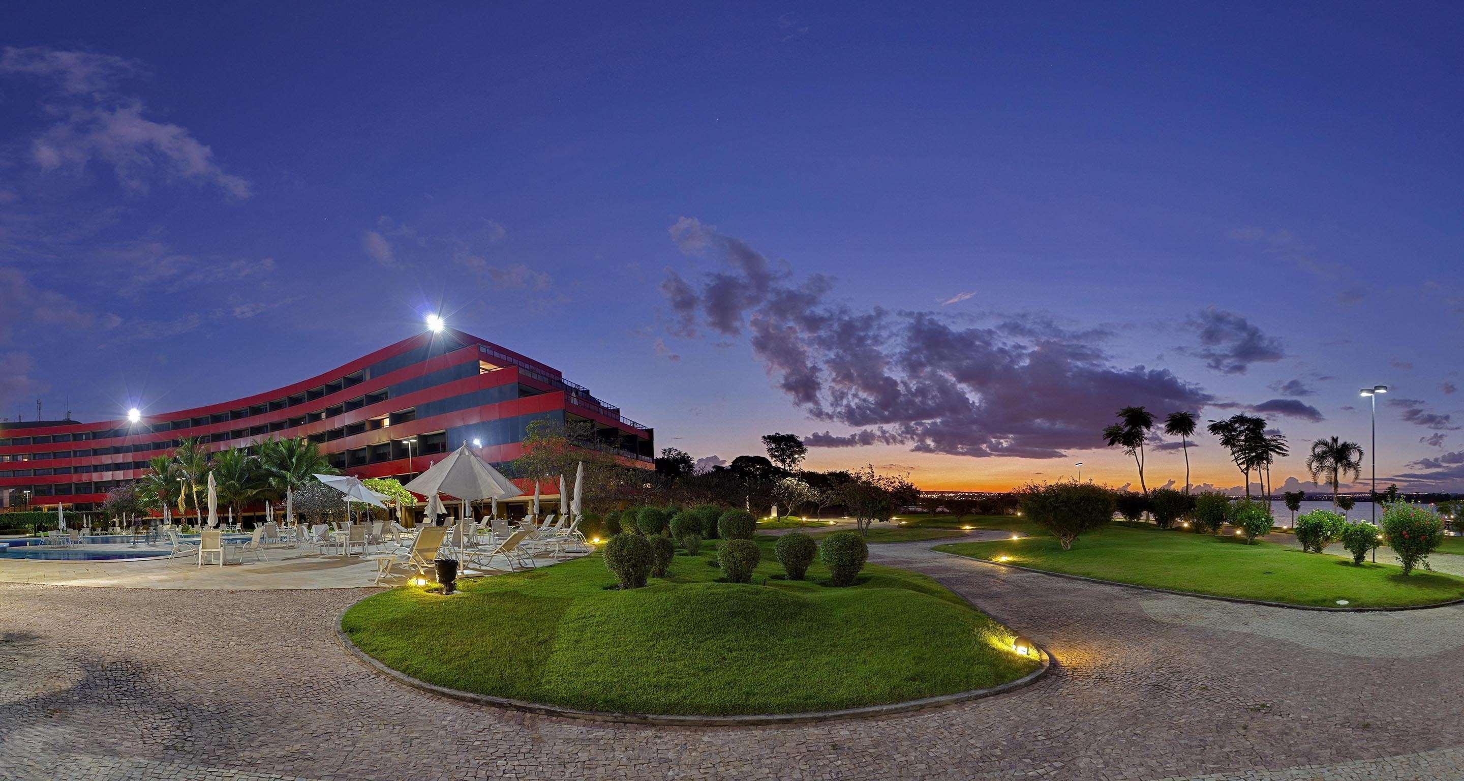 Отель Royal Tulip Brasilia Alvorada Экстерьер фото The photo depicts a modern building, possibly a hotel or resort, with a striking red exterior. It is set in a landscaped area that features well-maintained greenery and ornamental plants. In the foreground, there are lounge areas with umbrellas, sugg