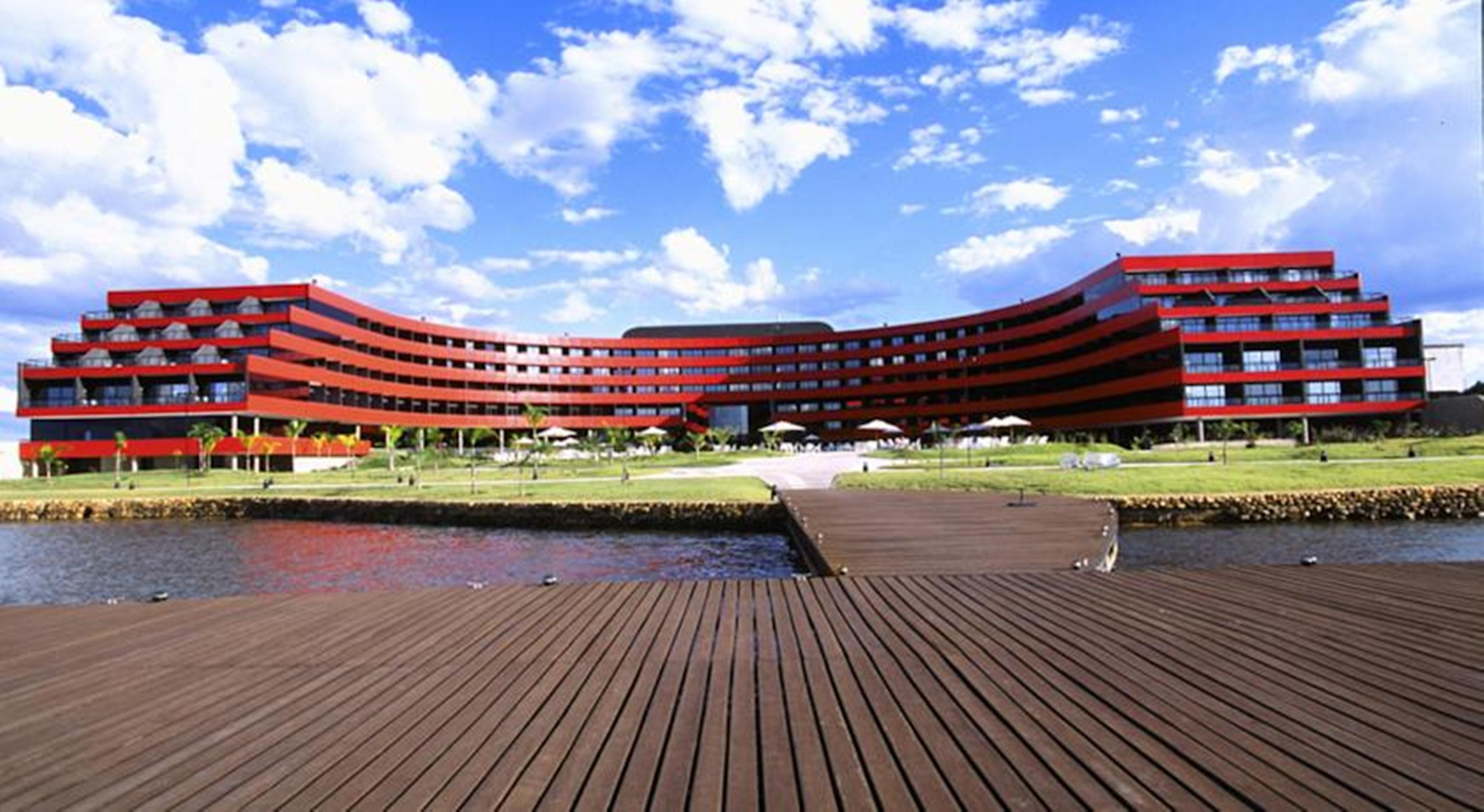 Отель Royal Tulip Brasilia Alvorada Экстерьер фото The image shows a modern, curved building with a striking red facade, situated near a body of water. The architecture features multiple levels with large windows that create a sleek and contemporary appearance. In front of the building, there is a wo