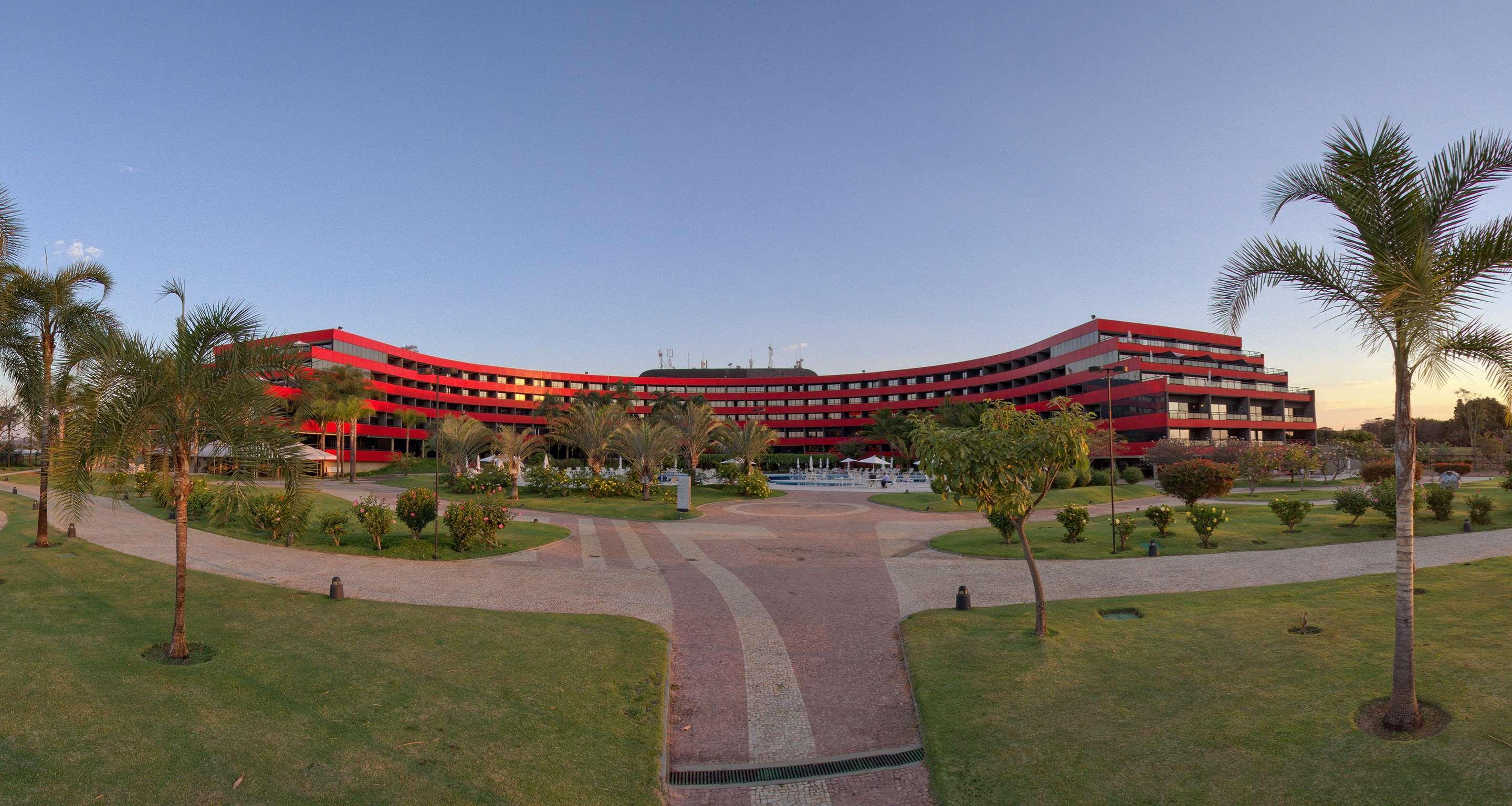 Отель Royal Tulip Brasilia Alvorada Экстерьер фото The photo shows a large, curved building with a vibrant red exterior. It appears to be a hotel or resort, surrounded by well-maintained gardens and green spaces. The landscape includes manicured lawns, small trees, and pathways, suggesting a pleasant