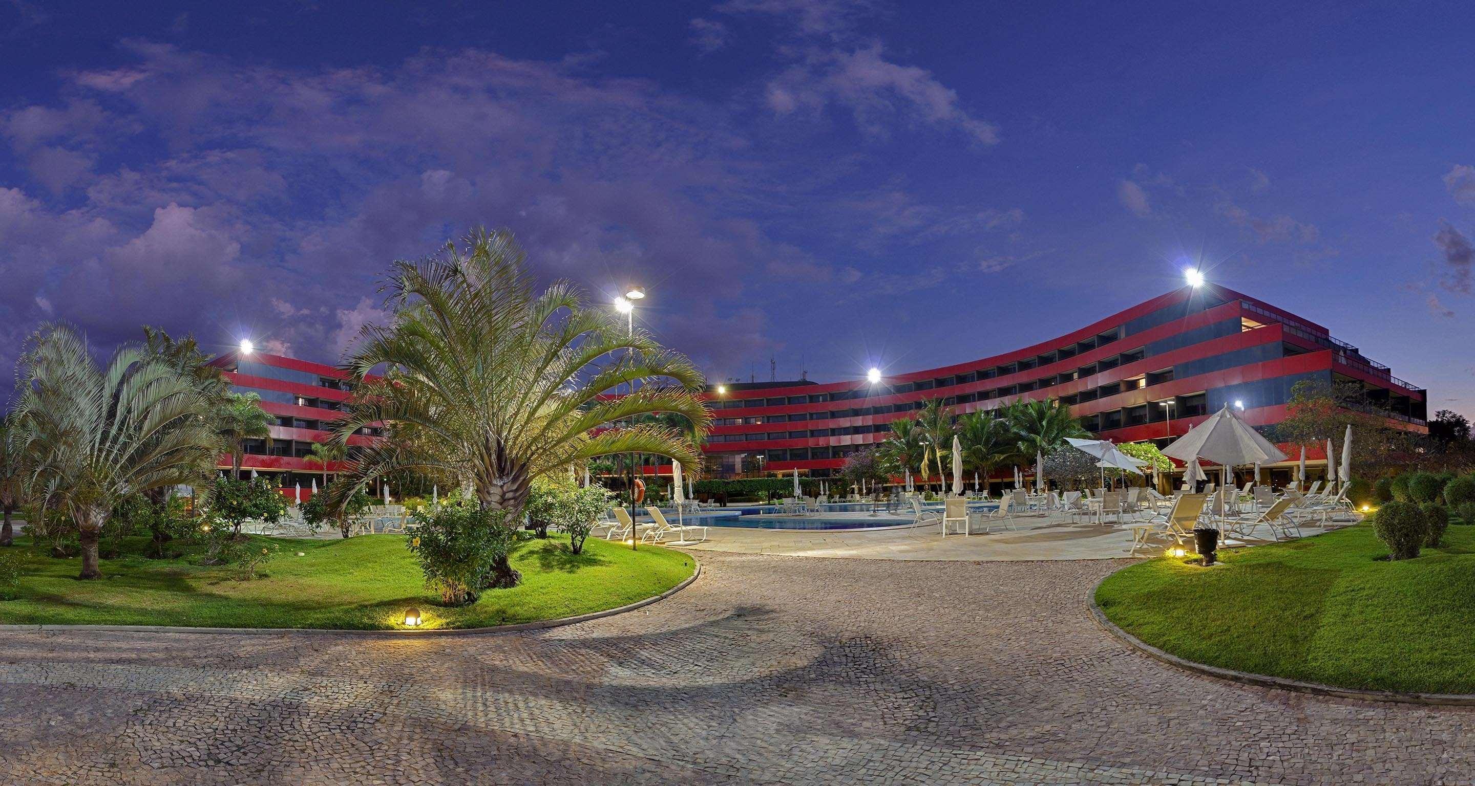 Отель Royal Tulip Brasilia Alvorada Экстерьер фото The photo shows a panoramic view of a hotel or resort complex, characterized by its modern architectural design with smooth, curved lines and a vibrant red color. It’s set in a landscaped area featuring palm trees and green grass. The surroundings in