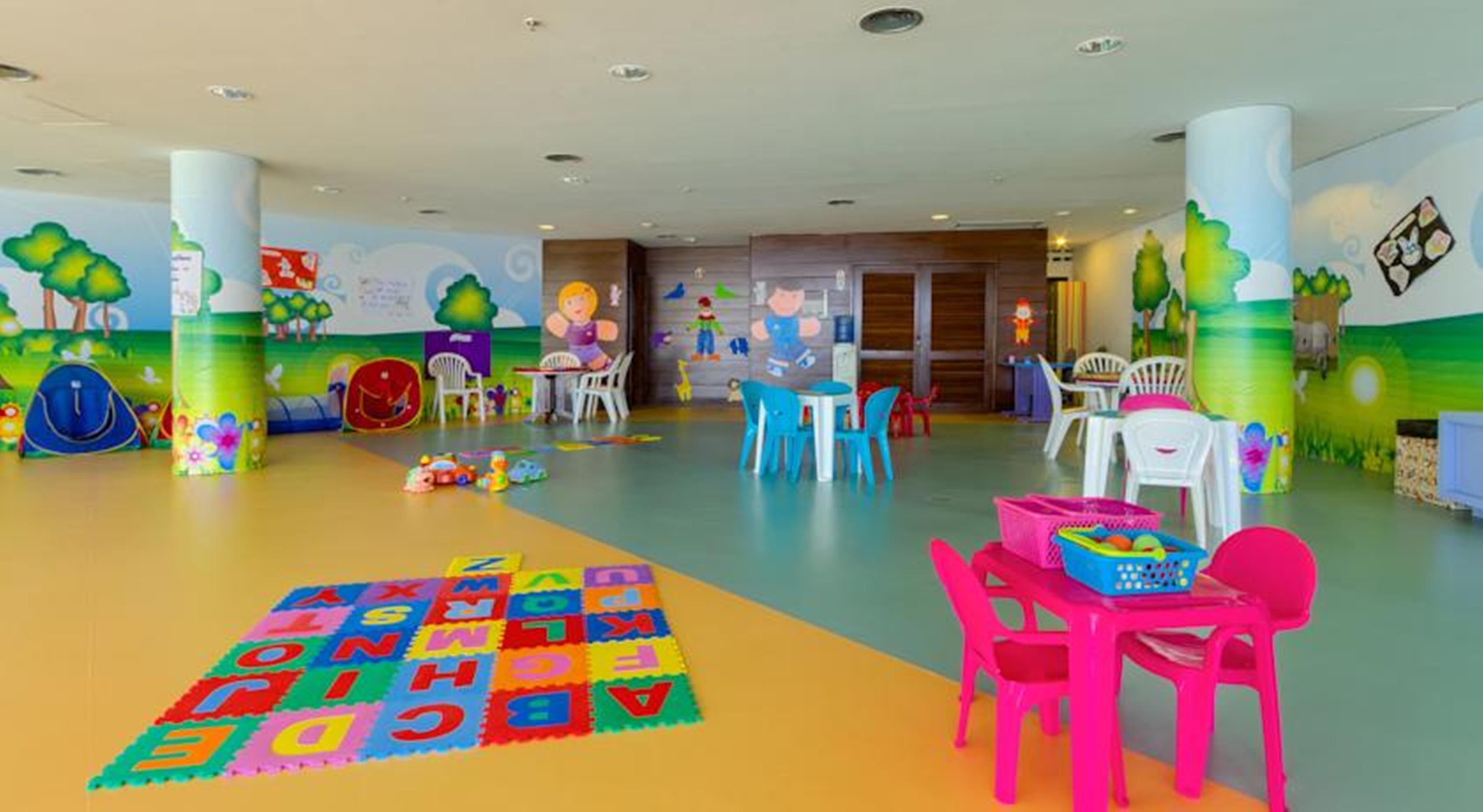 Отель Royal Tulip Brasilia Alvorada Экстерьер фото The photo shows a colorful and spacious children's playroom. The floor is divided into sections with different colors, and there is a large play mat featuring letters and numbers. 

In the room, there are several small, lightweight tables and chairs 
