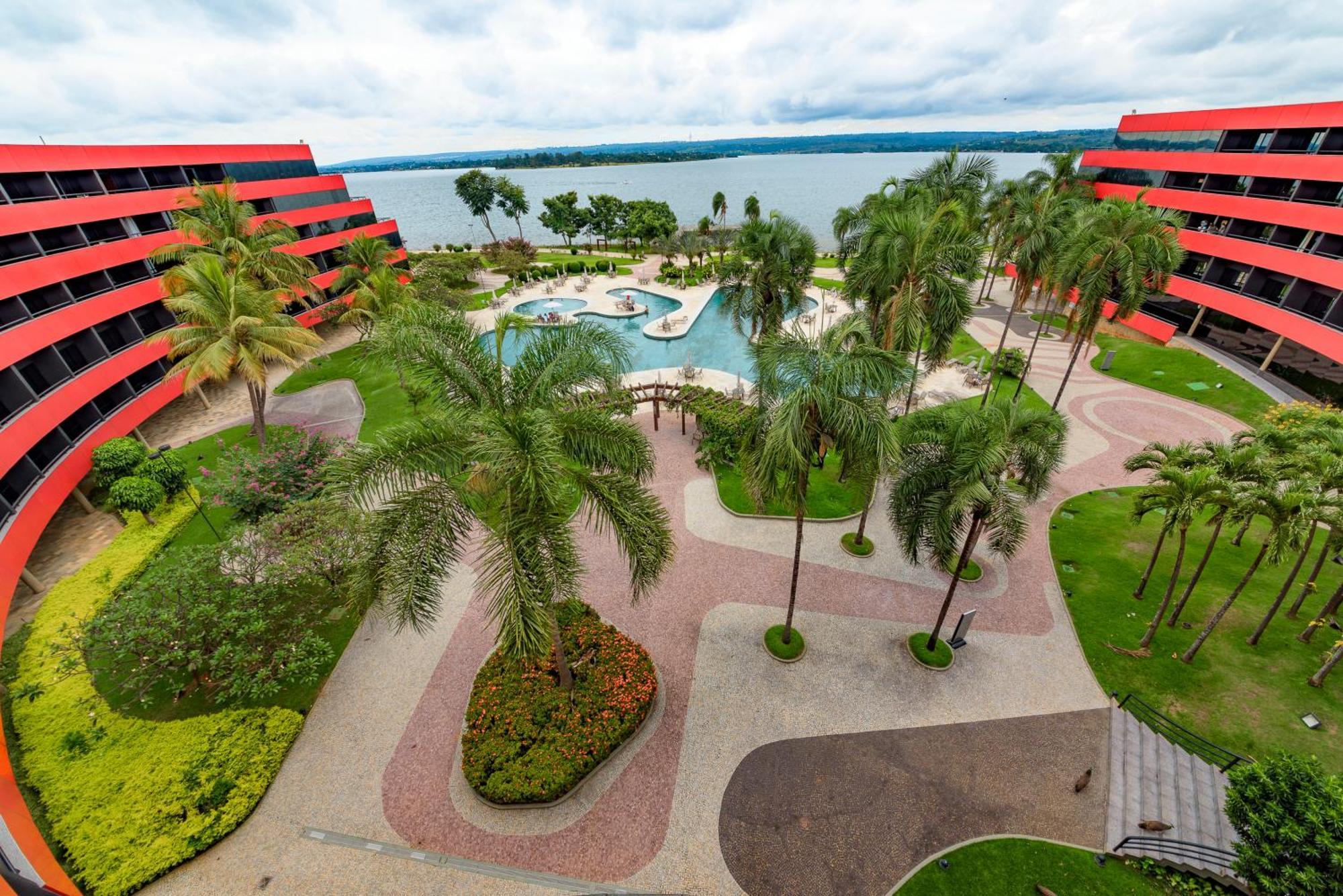 Отель Royal Tulip Brasilia Alvorada Экстерьер фото The photo shows a scenic view of a courtyard area in a residential complex. There are multiple red buildings surrounding a pool area, which is surrounded by palm trees and landscaped gardens. The pool features a curvy design, and there are pathways m