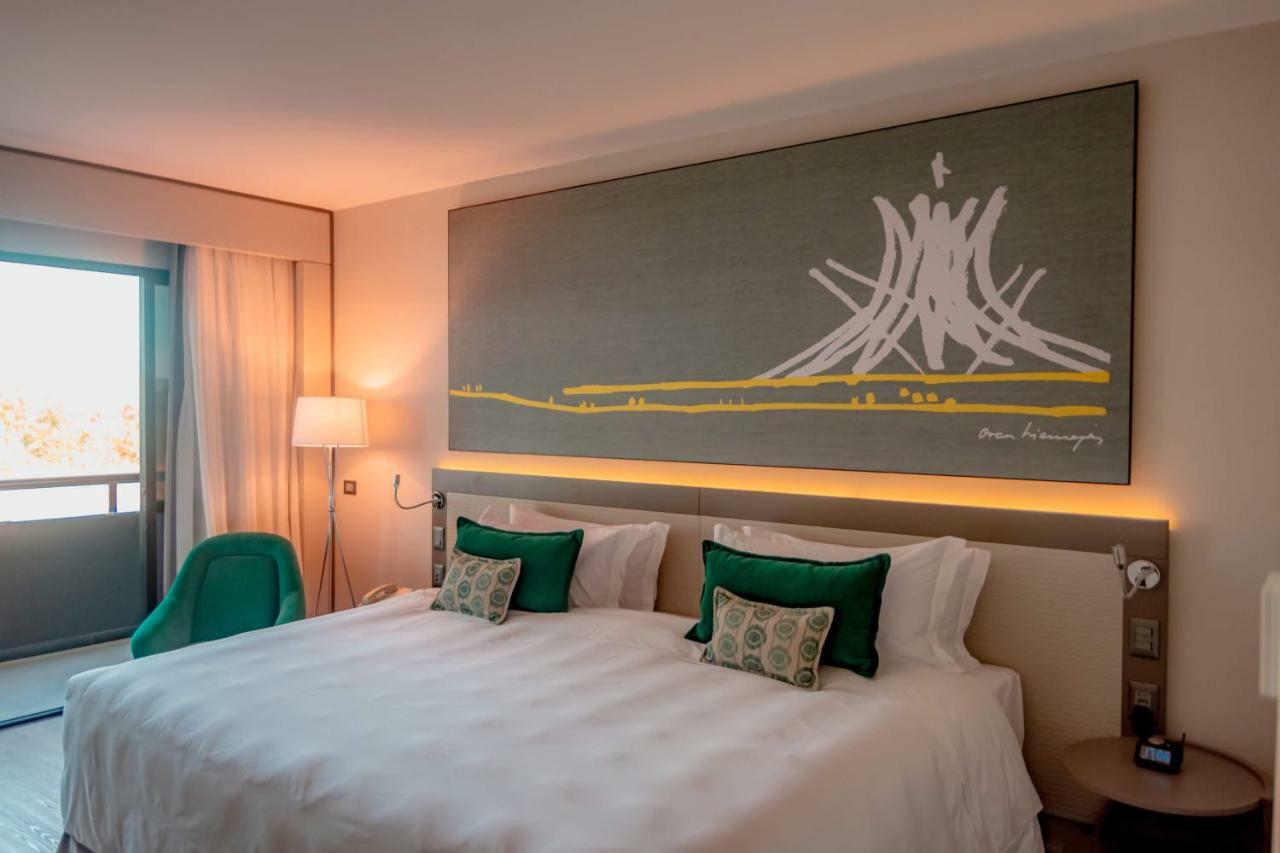 Отель Royal Tulip Brasilia Alvorada Экстерьер фото The photo shows a modern hotel room featuring a large bed with white bedding and two green accent pillows. Above the bed, there is a large piece of artwork depicting a stylized mountain or volcanic shape with yellow accents. The room has a contempora